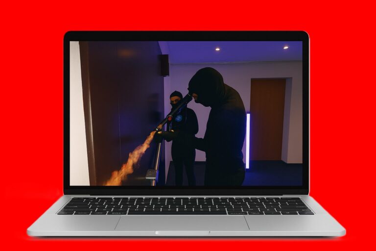 Laptop screen shows two masked individuals in dark clothing operating a flamethrower-like device emitting a flame in a dim room.
