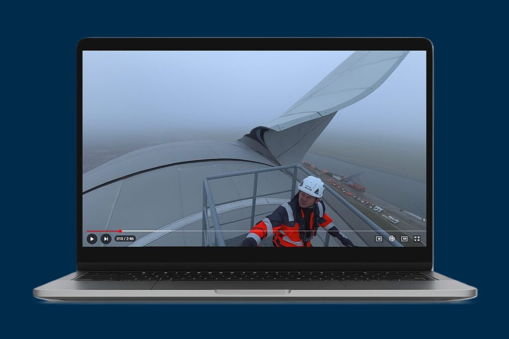 Video still: a maintenance worker in an orange safety jacket and white helmet stands on the wing of a large aircraft, overlooking the runway with fog and ground equipment visible below.